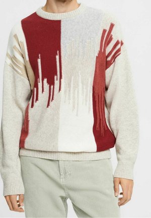 Pull avec des motifs verticaux abstraits rouges, beiges et blancs porté avec un pantalon en velours côtelé de couleur claire par une personne.