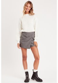 Pull blanc texturé avec des manches ballon, associé à une jupe portefeuille à carreaux gris et des bottines noires avec un accent en chaîne.