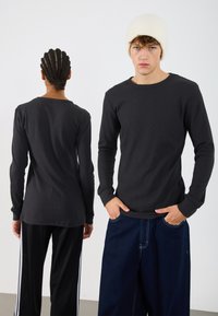 Camisola preta de mangas longas, justa, com textura canelada, combinada com jeans escuros largos. O modelo à esquerda tem cabelo trançado, enquanto o da direita usa um boné branco.