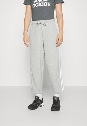 Person iført grå Adidas træningsbukser med hvide striber, en mørkegrå Adidas t-shirt, hvide sokker og sorte Adidas sneakers står på en grå gulv.