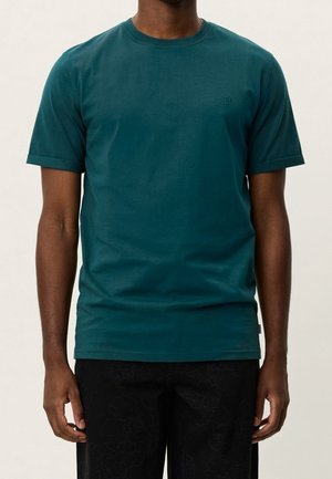 Personne portant un t-shirt uni bleu sarcelle à manches courtes et un pantalon noir, debout avec les bras détendus le long du corps devant un fond uni.