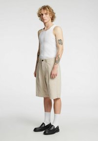 Biała bezrękawnikowa koszulka, beżowe plisowane szorty z kieszeniami, czarne mokasyny i białe skarpetki; krótkie włosy i widoczne tatuaże na ramionach.