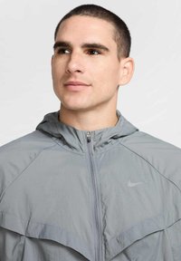 Giacca zip-up grigia chiara con cappuccio, caratterizzata da una texture liscia e un logo Nike impresso sul petto. Il design include dettagli di cuciture diagonali.