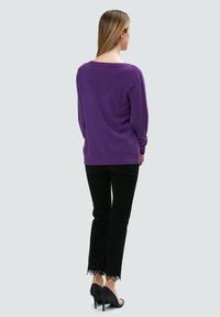 Lila Langarmpullover mit lockerer Passform, kombiniert mit schwarzen Jeans mit Spitzenverzierung und hohen schwarzen Absätzen, von hinten gezeigt.