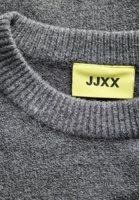 Gros plan sur l'encolure d'un pull en maille côtelée grise avec une étiquette de vêtement jaune vif affichant le nom de la marque JJXX en lettres noires.