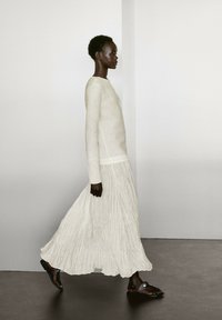 Helles, strukturiertes Creme-Kleid mit langen Ärmeln und plissiertem Rock; kombiniert mit dunklen Sandalen. Profilansicht vor einem minimalistischen Hintergrund.