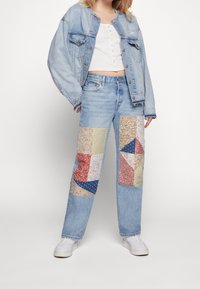 Denimová bunda a džíny s barevným patchworkovým aplikováním v různých květinových a geometrických vzorech. Bílé tenisky dotvářejí celý outfit.