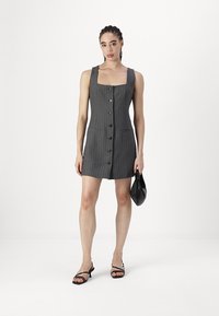 Šedé pruhované šaty se čtvercovým výstřihem, předním zapínáním na knoflíky a dvěma kapsami. Modelka drží malou černou kabelku a má na sobě sandály na řemínky.