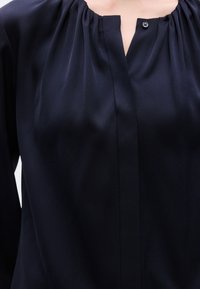 Μπλούζα σε ναυτικό μπλε χρώμα με απαλή, λεία υφή, με λαιμόκοψη με πιέτες, λεπτομέρεια με κουμπιά στο στήθος και ευθεία ραφή μπροστά.