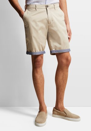 Beige Baumwollshorts mit umgeschlagenen Denim-Ärmeln, ausgestattet mit Vorderaschen und einem Knopfverschluss. Getragen mit beige Rutschschuhen.