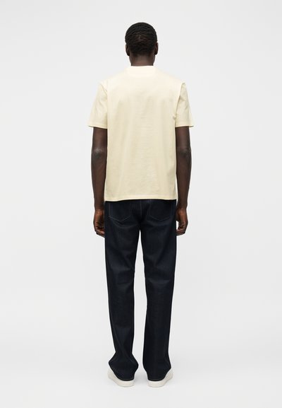 Άνδρας στέκεται γυρισμένος από την πλάτη, φοράει ένα απλό ανοιχτό μπεζ κοντομάνικο μπλουζάκι, σκούρο μπλε τζιν και λευκά αθλητικά παπούτσια, σε απλό φόντο.