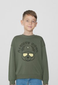 Zelená bavlněná mikina s potiskem medvěda v brýlích a čepici, s nápisem „VŽDY COOL“ nahoře v černé barvě.