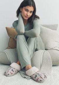 Graue Hausschuhe mit weißem, flauschigem Fellbesatz mit Hundemuster. Das Modell trägt ein hellgrünes Outfit und sitzt auf einer grau strukturierten Decke.