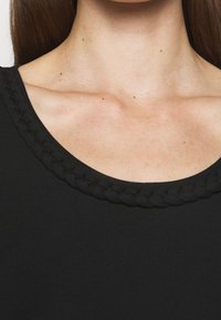 Svart topp med rund halsringning och flätad trimdetalj. Tyget verkar slätt och har en figurnära design.