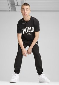 Czarna bawełniana koszulka z białym logo "PUMA", zestawiona z czarnymi dresami. Dodatki to srebrny łańcuszek i białe sneakersy.