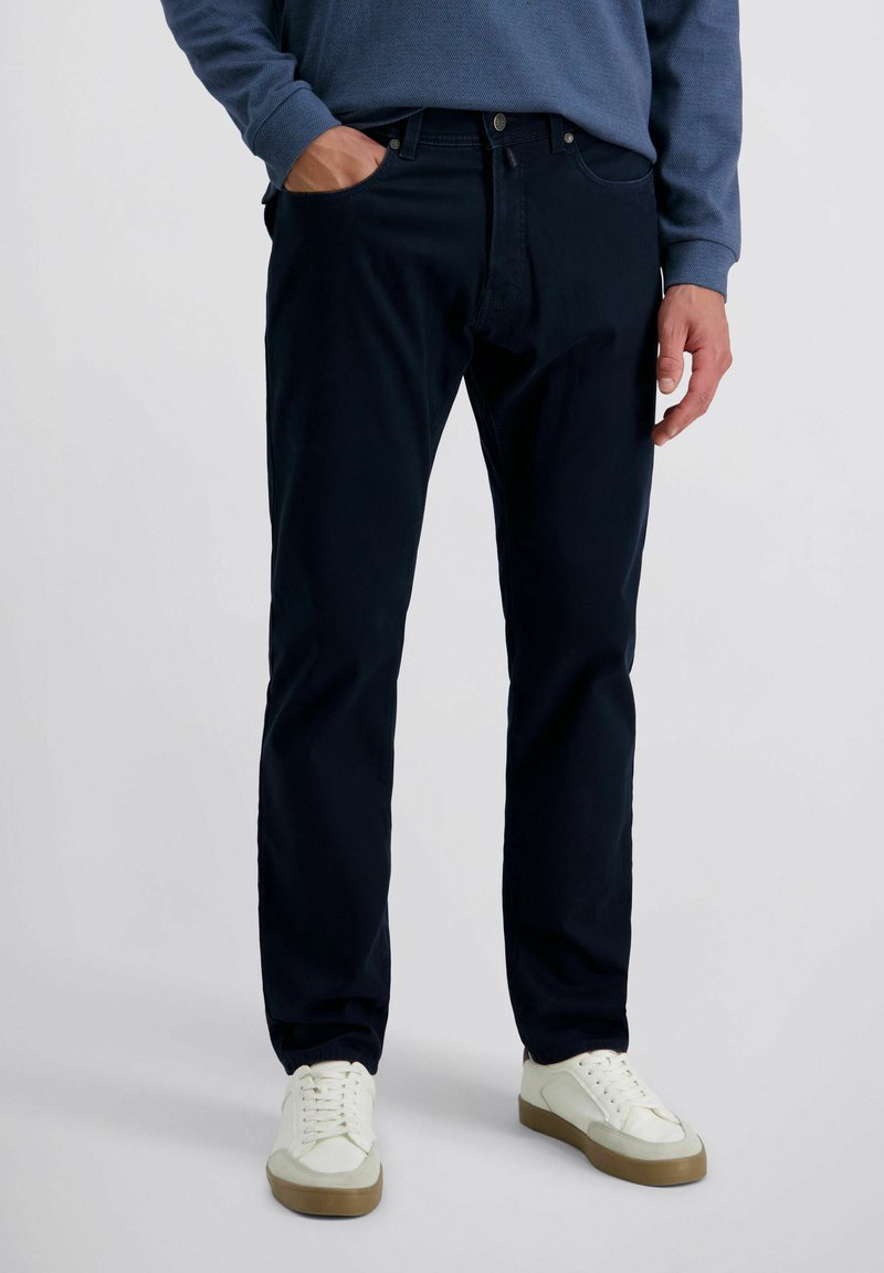 Mann trägt dunkelblaue gerade geschnittene Hose, blaues langärmliges Hemd und weiße Sneakers und steht mit einer Hand in der Tasche.