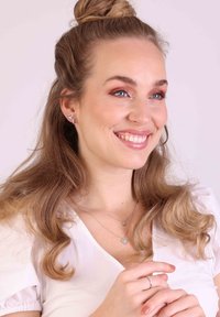 Vitrine de bijoux présentant des boucles d'oreilles en argent en forme de cœur et des colliers superposés. Le sujet a des cheveux ondulés et porte un haut blanc, mettant en valeur les accessoires.