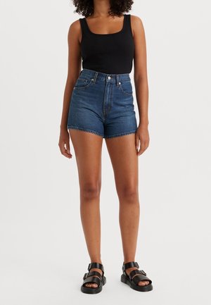 Vrouw met een zwart mouwloos topje, hoge taille blauwe denim shorts en zwarte sandalen met gesp, staand tegen een witte achtergrond.