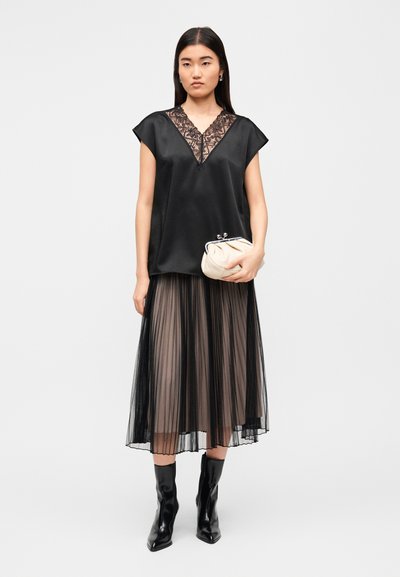 Μαύρο σατέν μπλουζάκι με δαντελένιο V-λαιμόκοψη, σε συνδυασμό με μαύρη πλισέ φούστα και μαύρα μπότες μέχρι το γόνατο, κρατώντας ένα κρεμ φάκελο.