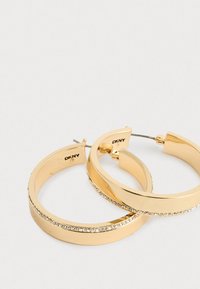 Cercei în stil huggie de culoare aurie, cu un rând de cristale mici și clare pe marginea exterioară, marcă DKNY, așezați pe o suprafață albă.