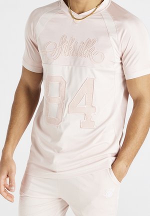 Hombre vistiendo camiseta deportiva SikSilk rosa claro con el número 84 y pantalones cortos a juego, una mano en el bolsillo, cadena de oro visible.