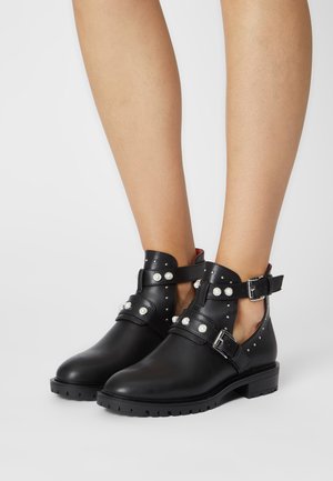 Bottes noires à la cheville avec des clous et des boucles argentés, portées sur des jambes nues, sur un fond clair uni.