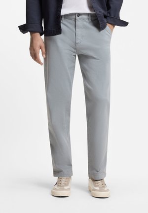 Voksen iført lysegrå bukser med lige ben, beige og hvide sneakers, hvid skjorte og mørk jakke med hænder delvist i lommerne.