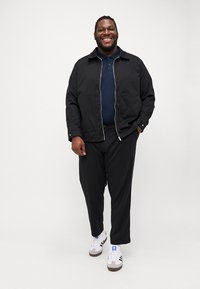 Svart lett jakke med glidelås, marineblå polo skjorte, svarte bukser og hvite joggesko, med mørkebrune detaljer.