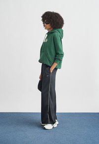 Zielona bluza z kapturem z przednią kieszenią, czarne spodnie zwężane z białymi bocznymi paskami oraz białe buty sportowe z zielonymi akcentami. Osoba nosi okulary przeciwsłoneczne.