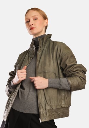 Olivově zelená kožená bomber bunda s vysokým límcem, zipem vpředu a bočními kapsami, nošená přes šedý svetr. Měkká textura, uvolněný střih.