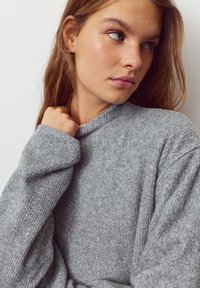 Pulover gri cu rețea răsucită, decolteu înalt și mâneci lungi, cu un tricot texturat și croială relaxată, expus pe un fundal neutru.