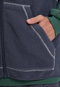 Hand med tummen i fickan på en strukturerad mörkblå fleecejacka med synlig vit söm på dragkedjan som går över en grön ärm.