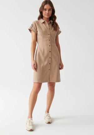 Abito camicia taupe con maniche corte, chiusura a bottoni e dettagli di cucitura sottili. Abbinato a sneakers bianche. Tessuto liscio e leggero.