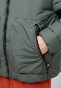 Giacca a vento in verde scuro, con cerniera frontale, tasche laterali con zip e una texture morbida e trapuntata.