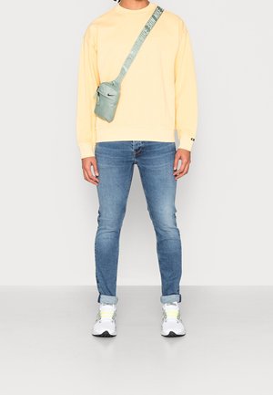 Gelber Pullover mit gerippten Bündchen, blaue Slim-Fit-Jeans und weiße Sneaker mit grünen Akzenten. Kleine mintgrüne Umhängetasche.