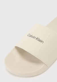 Beige glidesandal med en teksturert stropp med preget "Calvin Klein"-logo. Glatt fotseng og avrundet såledesign.