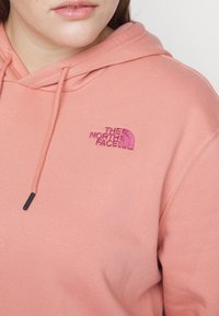 Gros plan sur une personne portant un sweat à capuche rose avec le logo "The North Face" brodé en rose plus foncé sur le côté gauche de la poitrine.