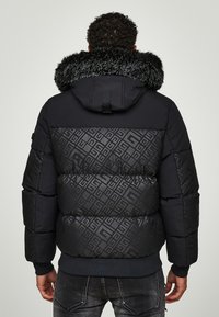 Giacca a vento nera con design trapuntato e motivo geometrico, cappuccio foderato in pelliccia e polsini a coste, mostrata da dietro.