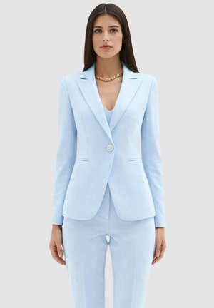 Donna che indossa un blazer azzurro chiaro aderente e pantaloni abbinati, accessoriata con una collana a catena dorata, in piedi davanti a uno sfondo uniforme.