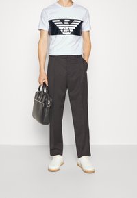 Man wearing white logo t-shirt, dark trousers, white sneakers, holding a black leather briefcase bag, standing against a white background.