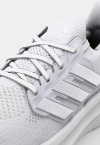 Weiße Sportschuhe mit einem strukturierten Strick-Obermaterial, flachen Schnürsenkeln und drei markanten weißen Streifen an der Seite. Mit einer gepolsterten Sohle.