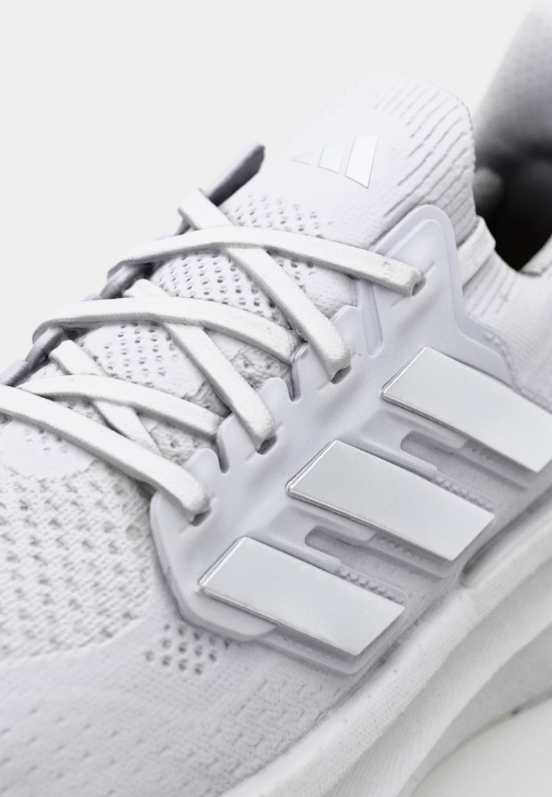 Weiße Sportschuhe mit einem strukturierten Strick-Obermaterial, flachen Schnürsenkeln und drei markanten weißen Streifen an der Seite. Mit einer gepolsterten Sohle.