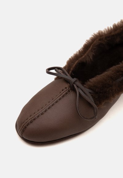 Gros plan d'une pantoufle mocassin marron avec une doublure douce et duveteuse et un nœud en cuir noué sur le dessus.