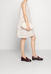 Beige ärmelloses Kleid mit strukturiertem Muster, knielanger Saum, kombiniert mit burgunderfarbenen Loafern mit goldenen Akzenten und einer cremefarbenen Clutch.