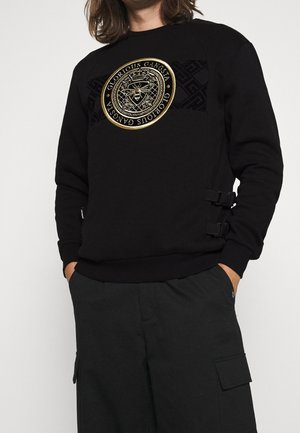Sudadera negra con un emblema redondo dorado con detalles intrincados, paneles negros con estampado en los hombros y hebillas en los lados.