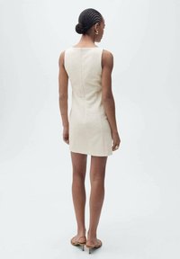 Robe courte crème avec une silhouette ajustée, design sans manches, tissu texturé et fermeture éclair au dos. Elle présente également un style minimaliste et une couleur neutre.