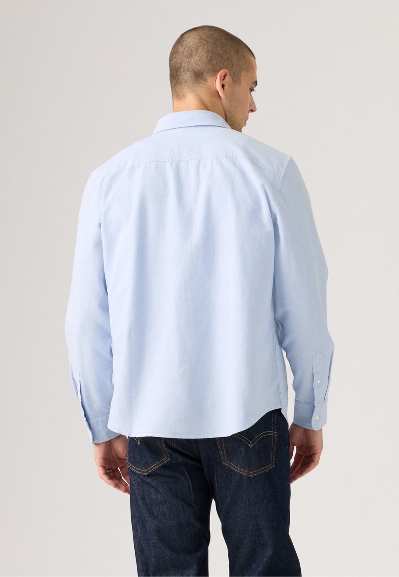 Light blue long-sleeve shirt with a button-down collar, back yoke, and curved hem, paired with dark denim jeans.