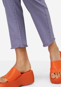 Sandalias de plataforma naranjas con una tira ancha, combinadas con pantalones de mezclilla morados recortados con dobladillos deshilachados y un diseño de pierna recta.