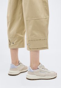 Κάτω άκρα με μπεζ παντελόνια καμπάνα που φτάνουν μέχρι τον αστράγαλο με μανσέτες με κουμπιά και μπεζ αθλητικά παπούτσια Lacoste με λευκές σόλες.