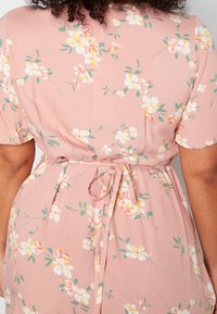 Kwiatowa sukienka w odcieniu delikatnego różu, pokryta białymi i żółtymi kwiatami, z krótkimi rękawami, ze zwężoną talią i wiązaniem oraz gładką fakturą.